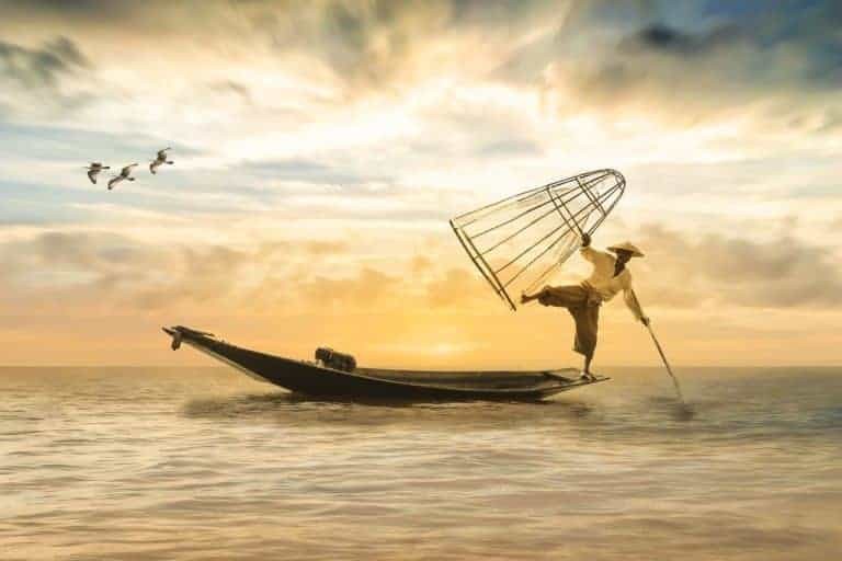 Рыбак, балансирующей на краю лодки, держащий ловушки для рыбы над головой и шест, упирающийся в воду